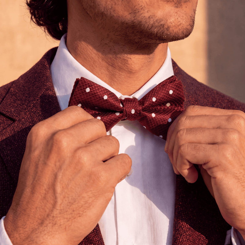 A close-up of a man adjusting a burgundy bow tie with white polka dots over a white collared shirt and a textured burgundy blazer.